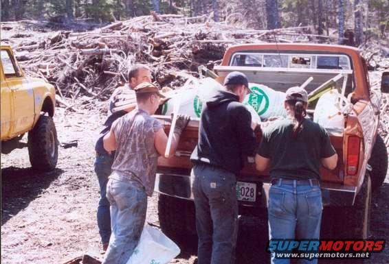 1976 Ford F-100 pickup truck with people loading bags into the bed, another yellow truck is partially visible on the left