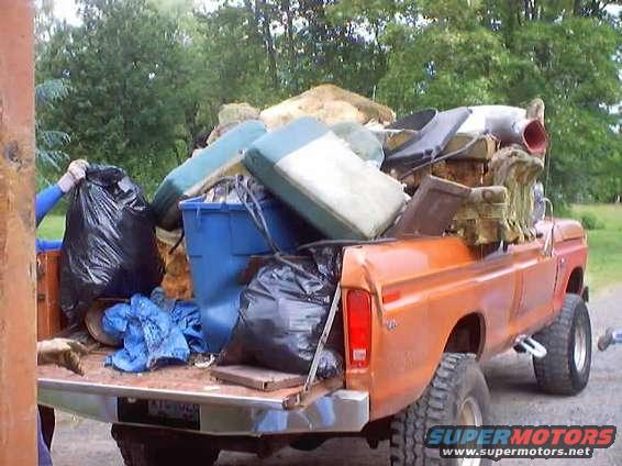 Orange 1976 Ford F-100 pickup truck loaded with trash bags and furniture, viewed from the rear