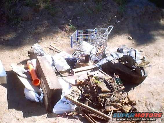 Pile of junk with a shopping cart, engine parts, wood, and bags on dirt ground