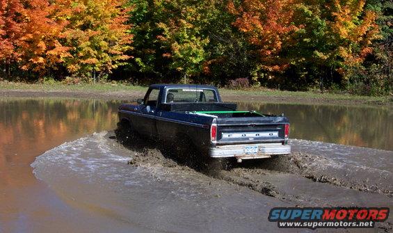 Dark blue Ford pickup driving through a muddy puddle with fall foliage in the background