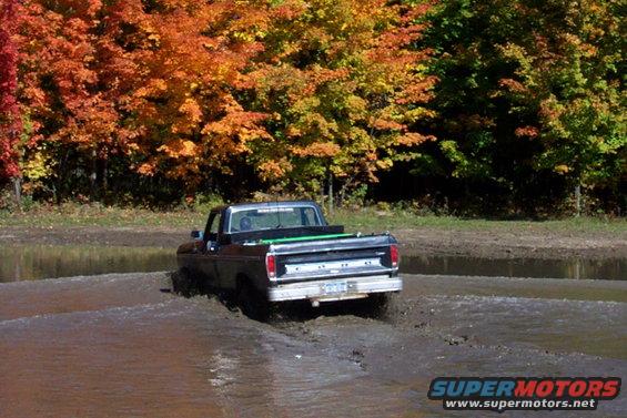 Dark blue Ford pickup truck driving through mud, autumn trees in background