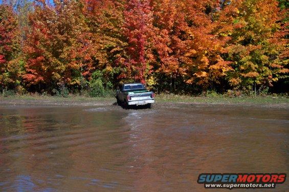 Green and white pickup truck driving through water with fall foliage in background