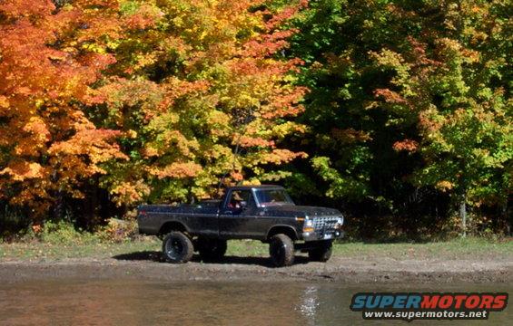 Black 1979 Ford F-Series pickup truck parked on muddy shore with fall foliage behind it