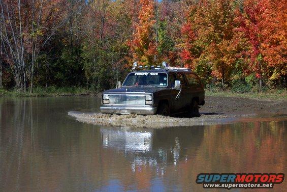 Dark SUV with chrome grille and roof rack driving through shallow water, autumn trees in background
