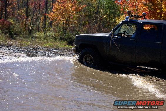 Dark pickup truck with off-road tires driving through shallow water, autumn trees in background