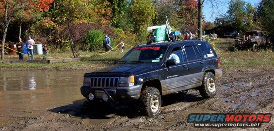 Black Jeep Grand Cherokee driving through mud with trees and people in the background. Another vehicle is visible in the distance