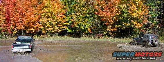 Pickup truck and Jeep driving through muddy water with autumn trees in background
