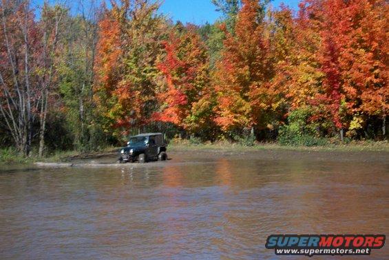 Jeep Wrangler driving through shallow water with fall foliage in background