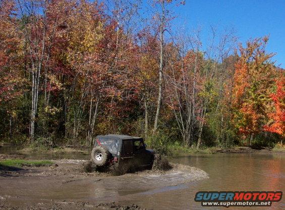Black Jeep Wrangler driving through mud, spare tire visible on rear