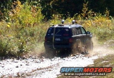 Dark colored Jeep Grand Cherokee driving through mud and water on a dirt trail. Has roof rack with lights