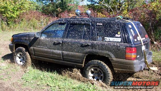 Mud-covered black Jeep Grand Cherokee driving through a muddy trail with trees in the background