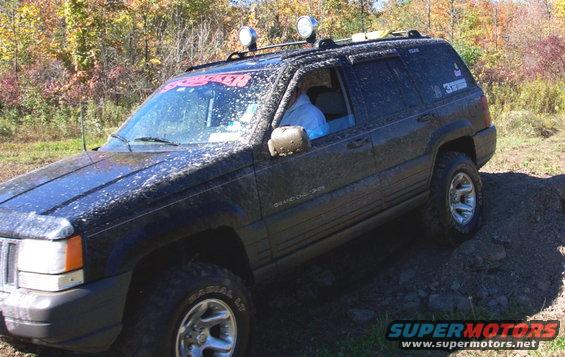 Black Jeep Grand Cherokee covered in mud, driving off-road with two lights on the roof rack