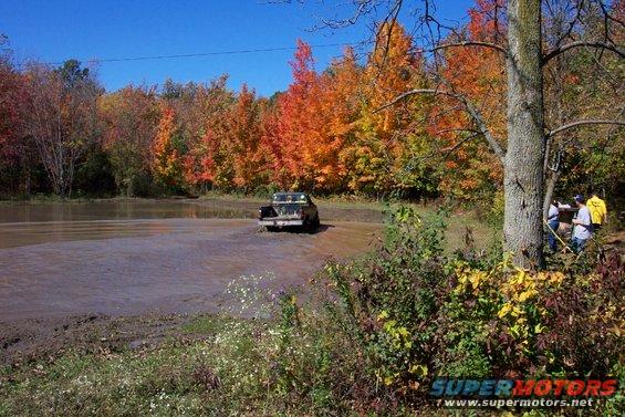 Truck driving through a muddy water crossing with fall foliage in the background