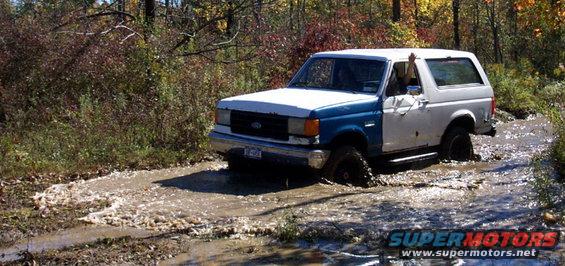Two-tone blue and white Ford Bronco stuck in mud, driver's arm raised