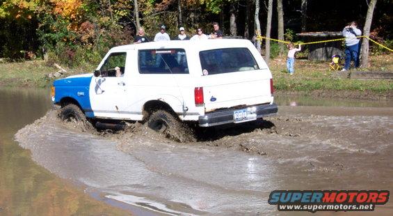 White and blue Ford Bronco driving through mud, rear view