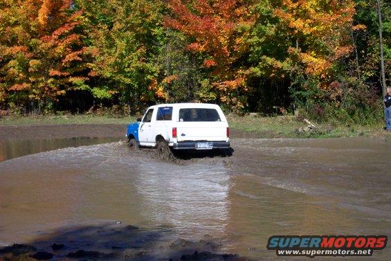 1979 Ford Bronco driving through a muddy puddle with fall foliage in the background