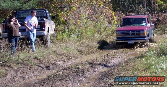 Two people stand by a lifted 1970s Ford Bronco as a red Ford Ranger drives through mud
