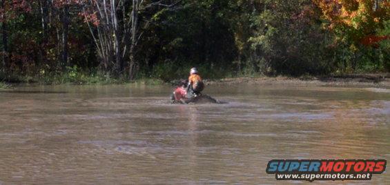 Person on an ATV rides through murky water with trees in the background