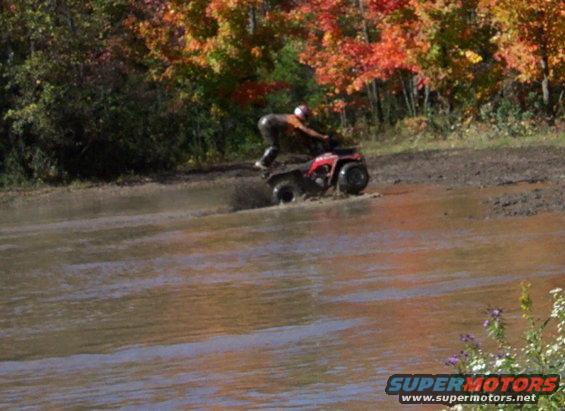 Red ATV with rider in muddy water, autumn trees in background