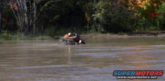 ATV with rider in orange shirt driving through muddy water with trees in background