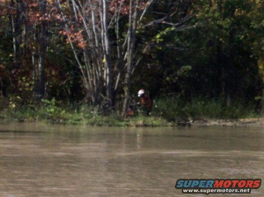 Person in orange jacket and white helmet using a chainsaw near trees by a body of water