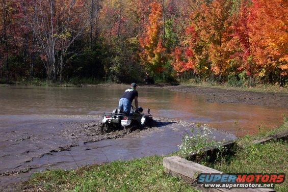 White all-terrain vehicle with a rider navigating through mud and water, with autumn trees in the background