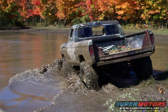 Muddy 1979 Ford F-Series pickup truck driving through shallow water and mud, autumn trees in background