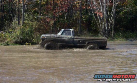 Muddy pickup truck driving through shallow water with trees in background