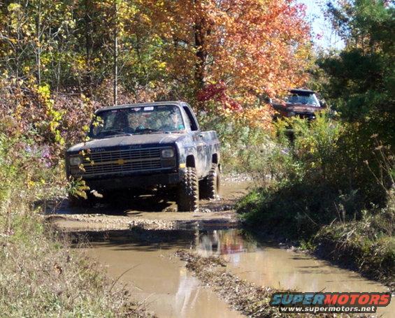 Two pickup trucks driving through a muddy trail in the woods with fall foliage