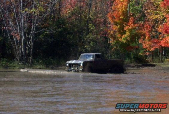 Mud-splattered 1970s Ford pickup truck driving through murky water with fall foliage in the background