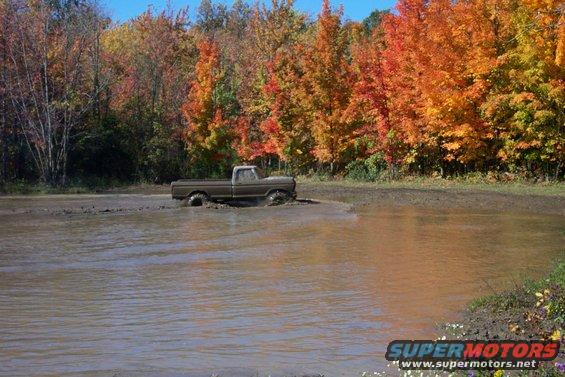 Brown pickup truck driving through mud with fall foliage in background
