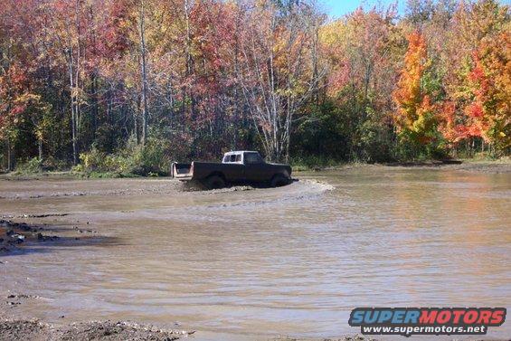 Dark colored pickup truck with white roof driving through a muddy area with fall foliage in the background