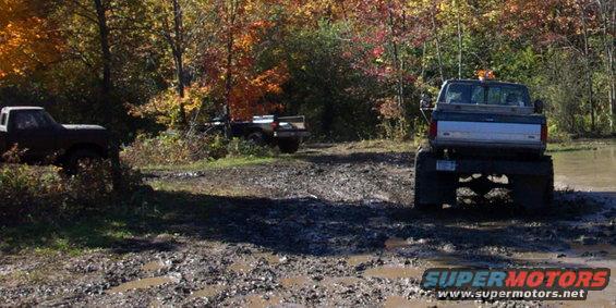 Three trucks driving through a muddy, wooded area during autumn