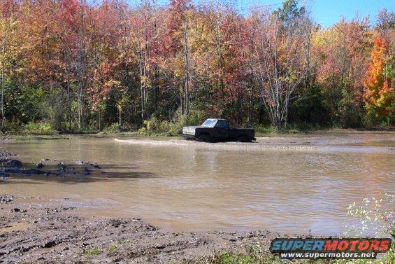 Black pickup truck driving through a muddy water hole with fall foliage in the background. Supermotors.net logo visible
