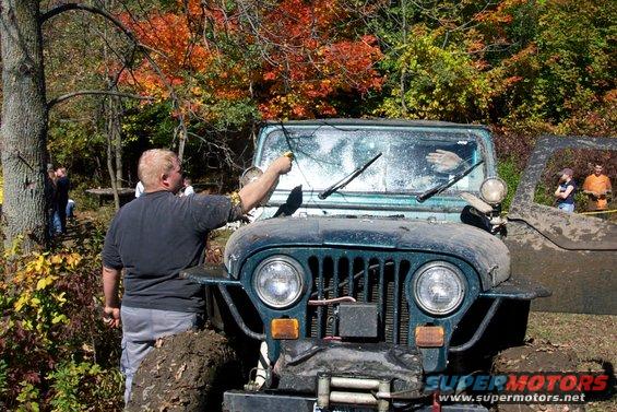 Dark green Jeep Wrangler stuck in mud, front view, with a person cleaning the windshield