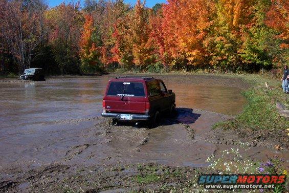 Red SUV driving through mud pit with fall foliage in background. Another dark SUV is visible in the distance
