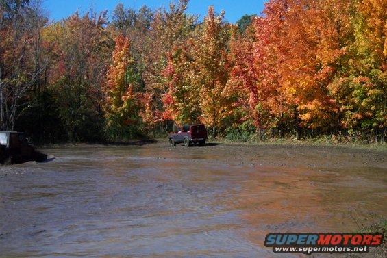 two SUVs parked in a muddy area with colorful fall trees in the background
