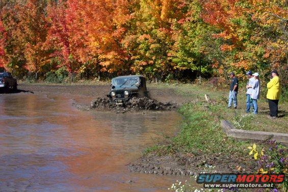 Green off-road vehicle splashing through mud with fall foliage in background