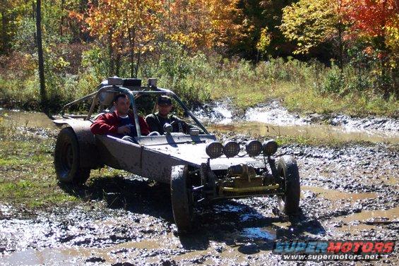 Two people in a buggy driving through mud and water with trees in the background