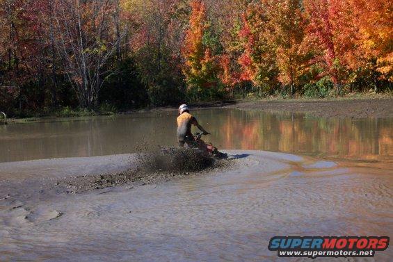 Person on ATV splashing through mud with fall foliage in background