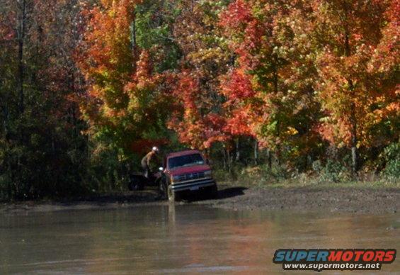 Red pickup truck partially submerged in mud, person on ATV nearby, autumn trees in background