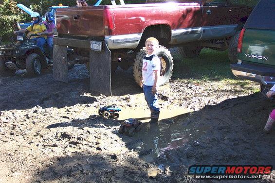 Kid in mud puddle with toy trucks, big red truck, green Yukon, and ATV in background
