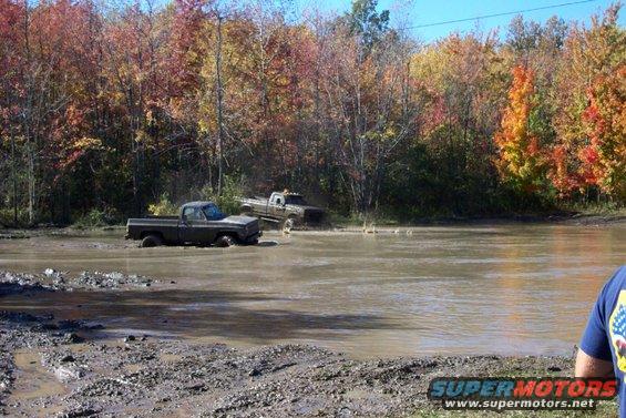Two trucks are stuck in a muddy water hole with fall foliage in the background