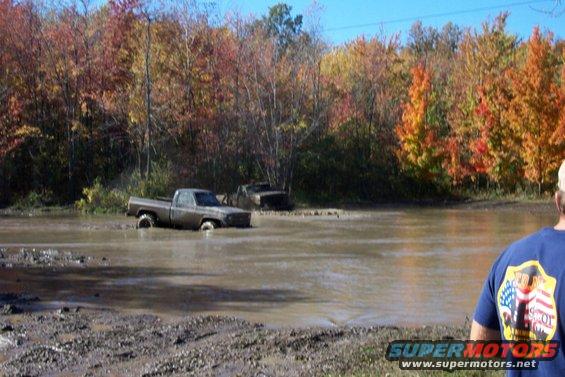 Two muddy trucks partially submerged in water with autumn trees in the background