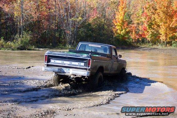 Black Ford pickup truck driving through mud with fall foliage in background. License plate visible