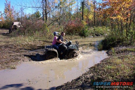 Green ATV with two riders splashing through a muddy puddle in a wooded area