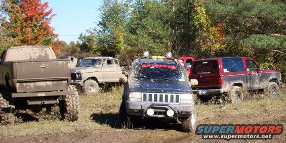Several muddy off-road vehicles parked in a field with trees in the background. One Jeep is in the foreground