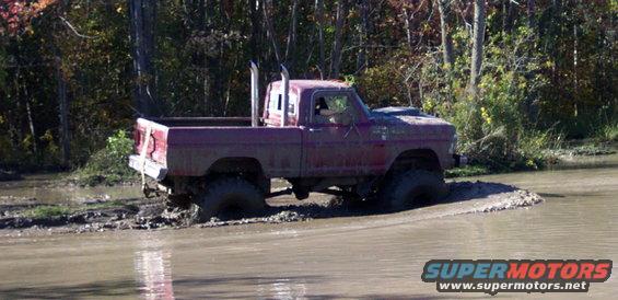 Mud-covered red pickup truck with dual exhaust stacks stuck in muddy water, trees in background
