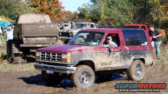 Red and black Ford Bronco II covered in mud, with other muddy trucks in the background