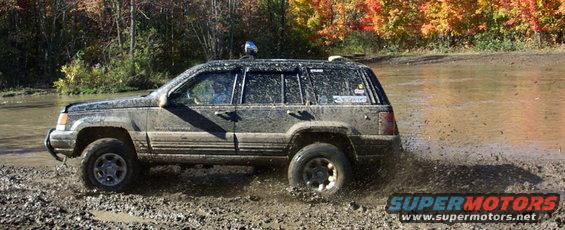 Muddy Jeep Grand Cherokee driving through a shallow water crossing with fall foliage in the background